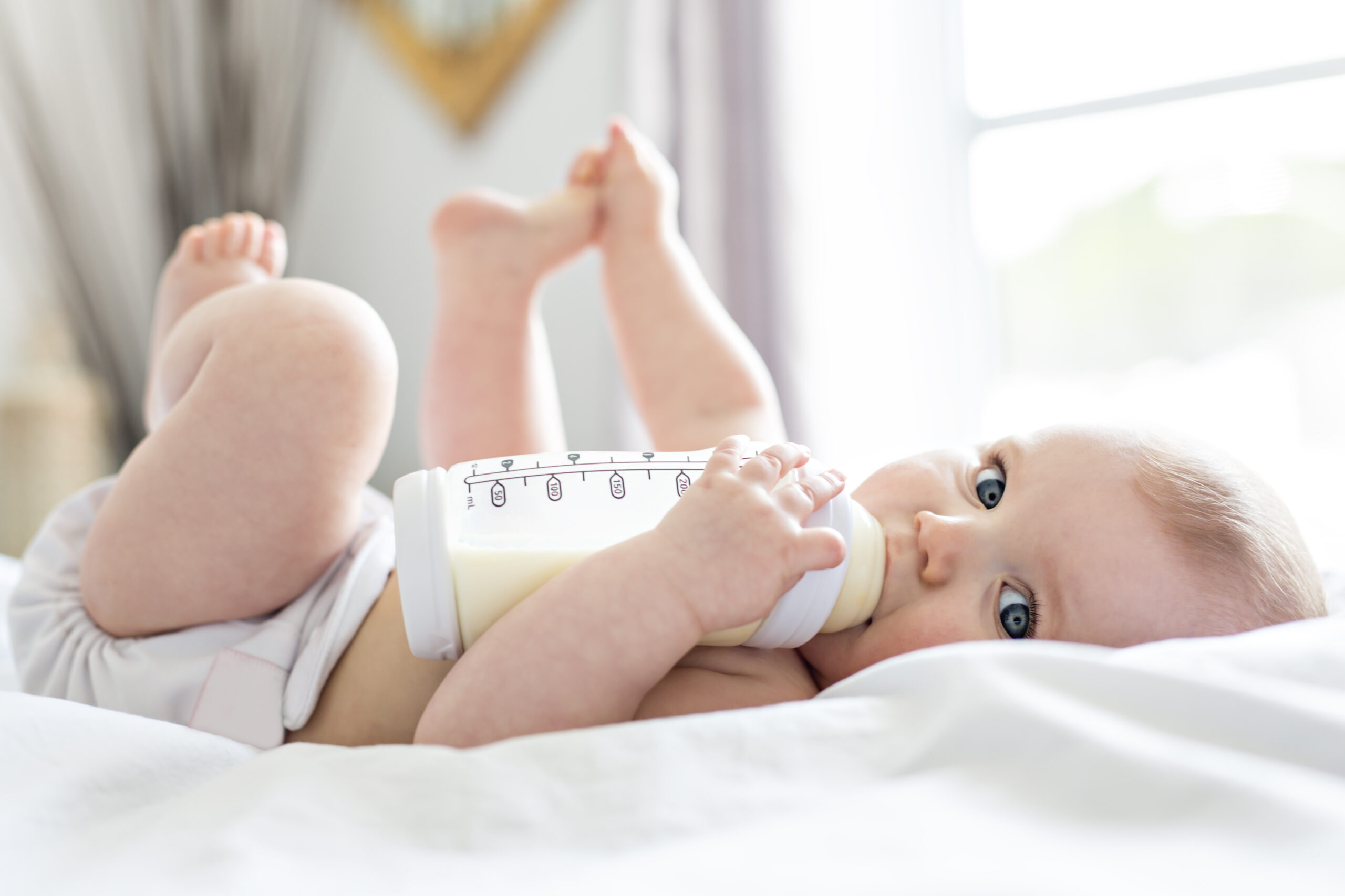 Pretty baby girl drinks water from bottle lying on bed. Child weared diaper in nursery room.